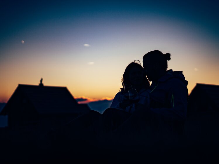 Paar sitzt im Gras im Sonnenuntergang vor der Almhütte