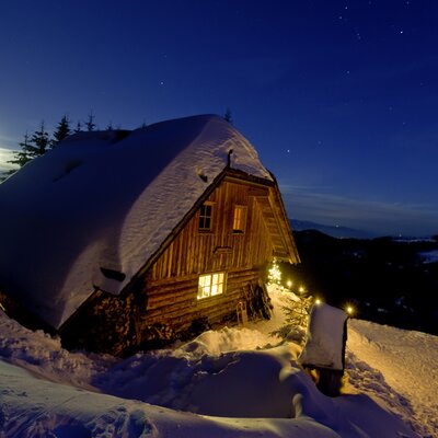 Kärntner Alm bei Nacht im Winter | © Urlaub am Bauernhof/ Tom Lamm Cozy wooden cabin nestled in snowy landscape, illuminated at night under starry sky. Serene winter scene. | © Urlaub am Bauernhof/ Tom Lamm