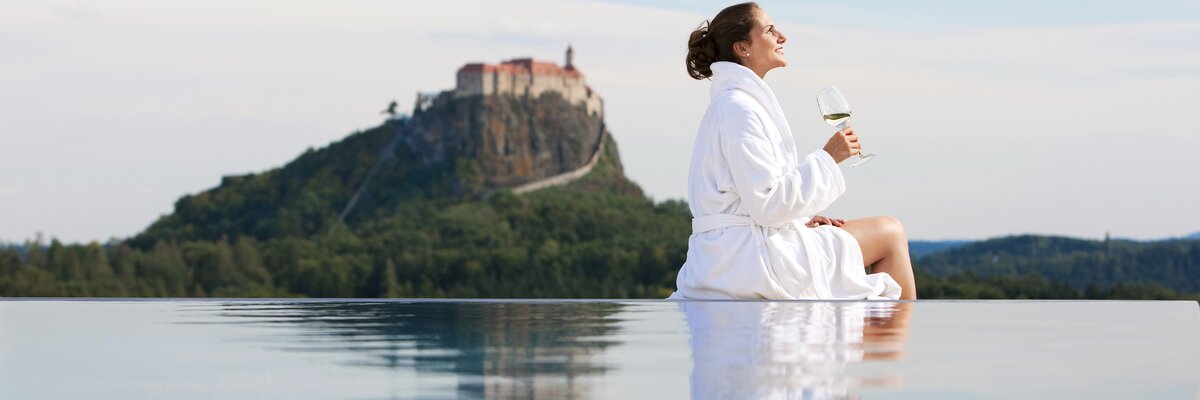 Eine Frau sitzt mit einem Weinglas am Beckenrand, im Hintergrund die Riegersburg, Thermen- & Vulkanland, Steiermark | © Thermen- & Vulkanland / Harald Eisenberger