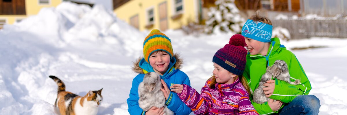 Drei Kinder sitzen im Schnee und streicheln Hasen und die Katze, Urlaubsregion Murau-Murtal, Steiermark | © Urlaub am Bauernhof Steiermark / Wolfgang Spekner