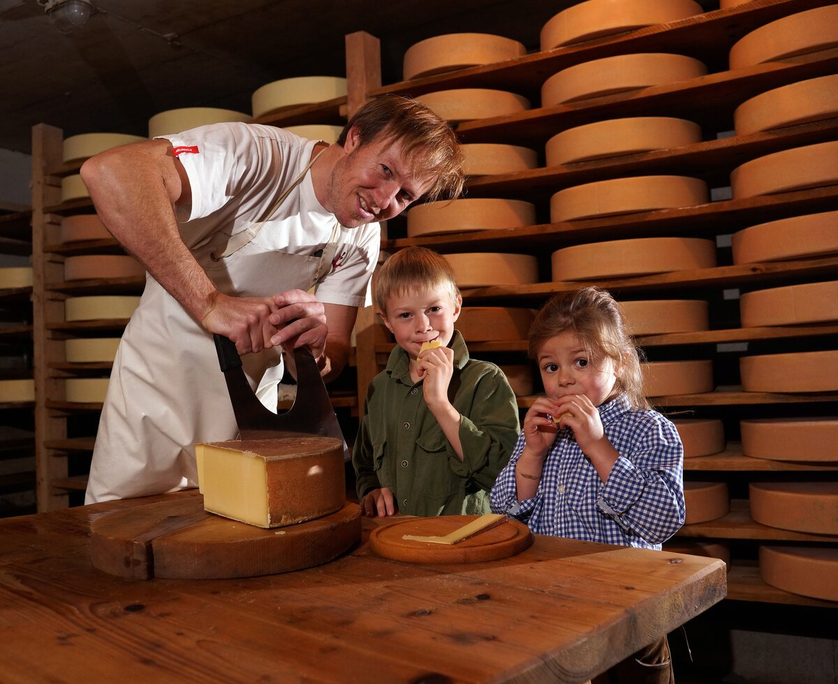 Senner schneidet einen Laib Käse an und Kinder kosten | © Urlaub am Bauernhof Vorarlberg / Ludwig Berchtold